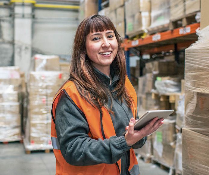 Warehouse worker smiles, holding tablet, wearing orange vest, standing near shelves of boxes.