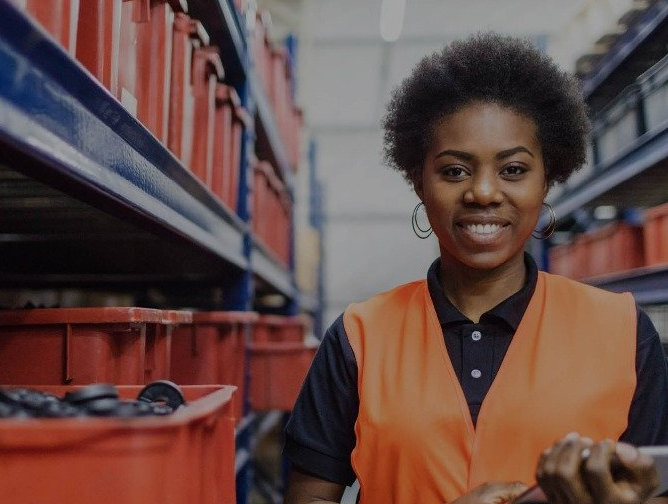 Woman in orange vest smiles in a warehouse, near shelves with red bins.