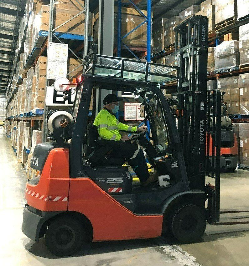 Forklift operator in a warehouse, wearing a face mask and safety vest, driving.