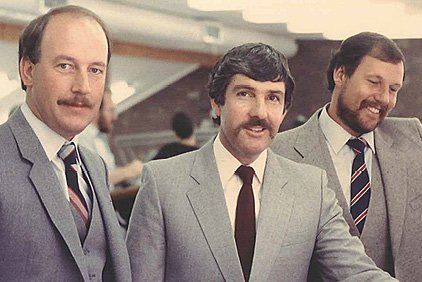 Three men in suits with mustaches smile, likely in an office setting.