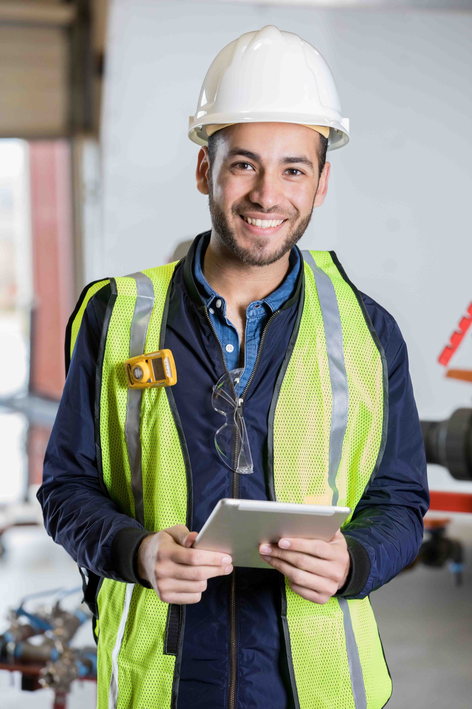 Construction worker in white hard hat and yellow vest holding tablet, smiling.