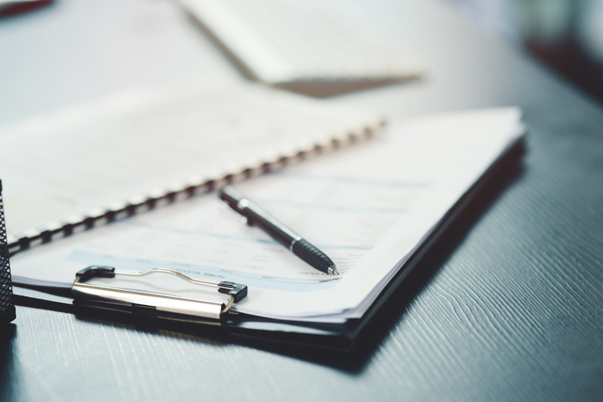 Clipboard with papers, spiral notebook, and pen on a dark wood desk.