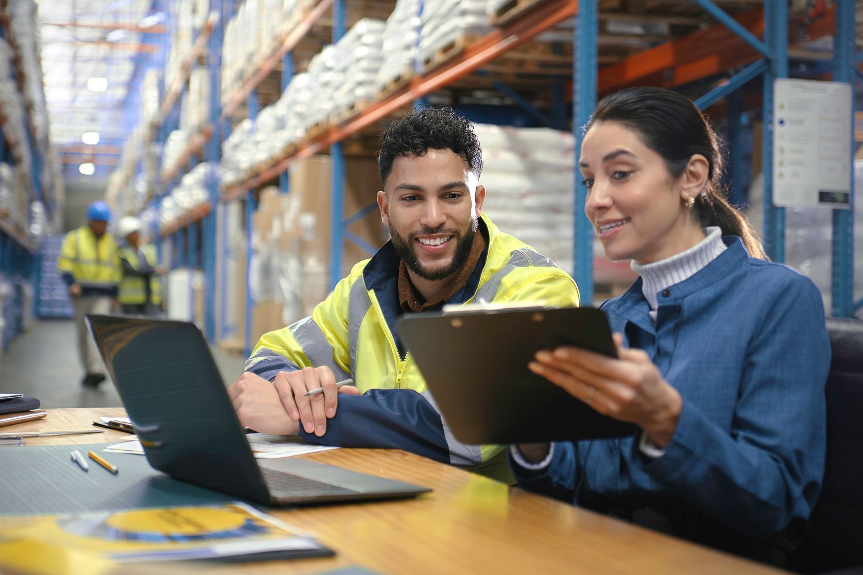 Man in safety vest and woman review documents at a desk in a warehouse.