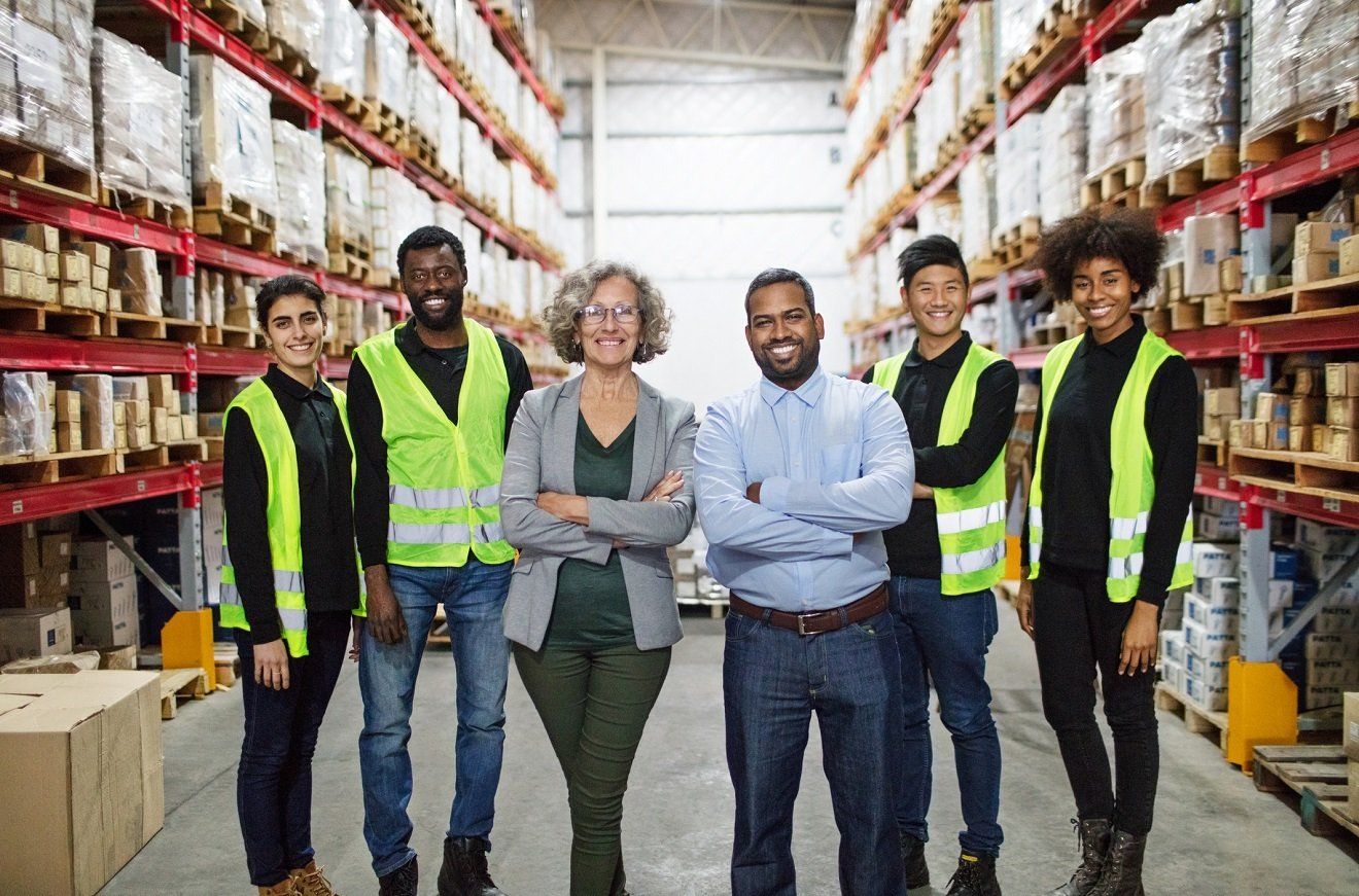 Warehouse team posing in front of shelves filled with boxes. All are smiling, wearing safety vests.
