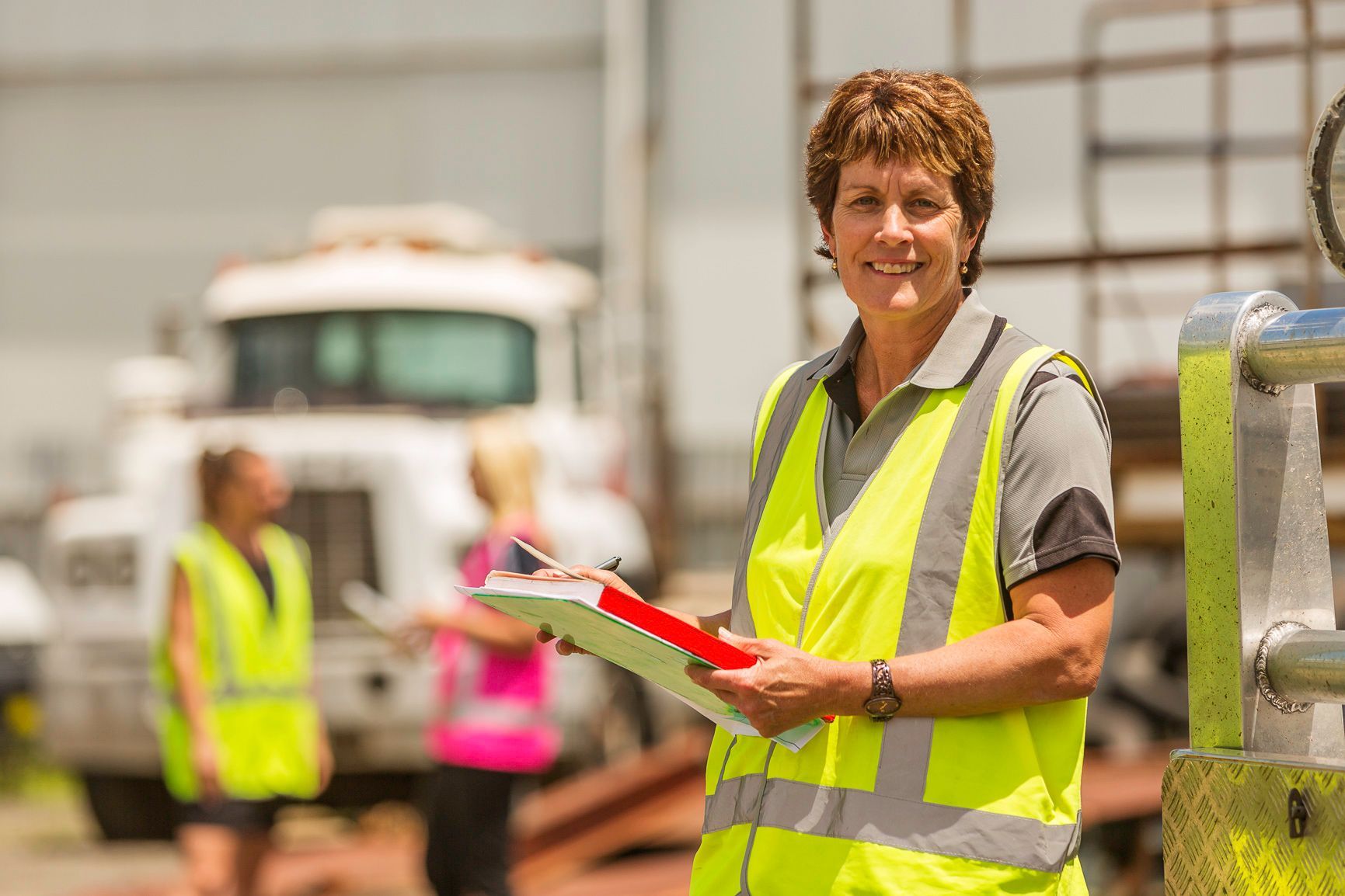 Woman in safety vest smiling, holding clipboard, in industrial setting, with trucks and other workers.