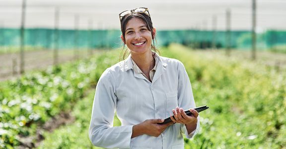 Woman in white lab coat smiles while holding tablet in a field.