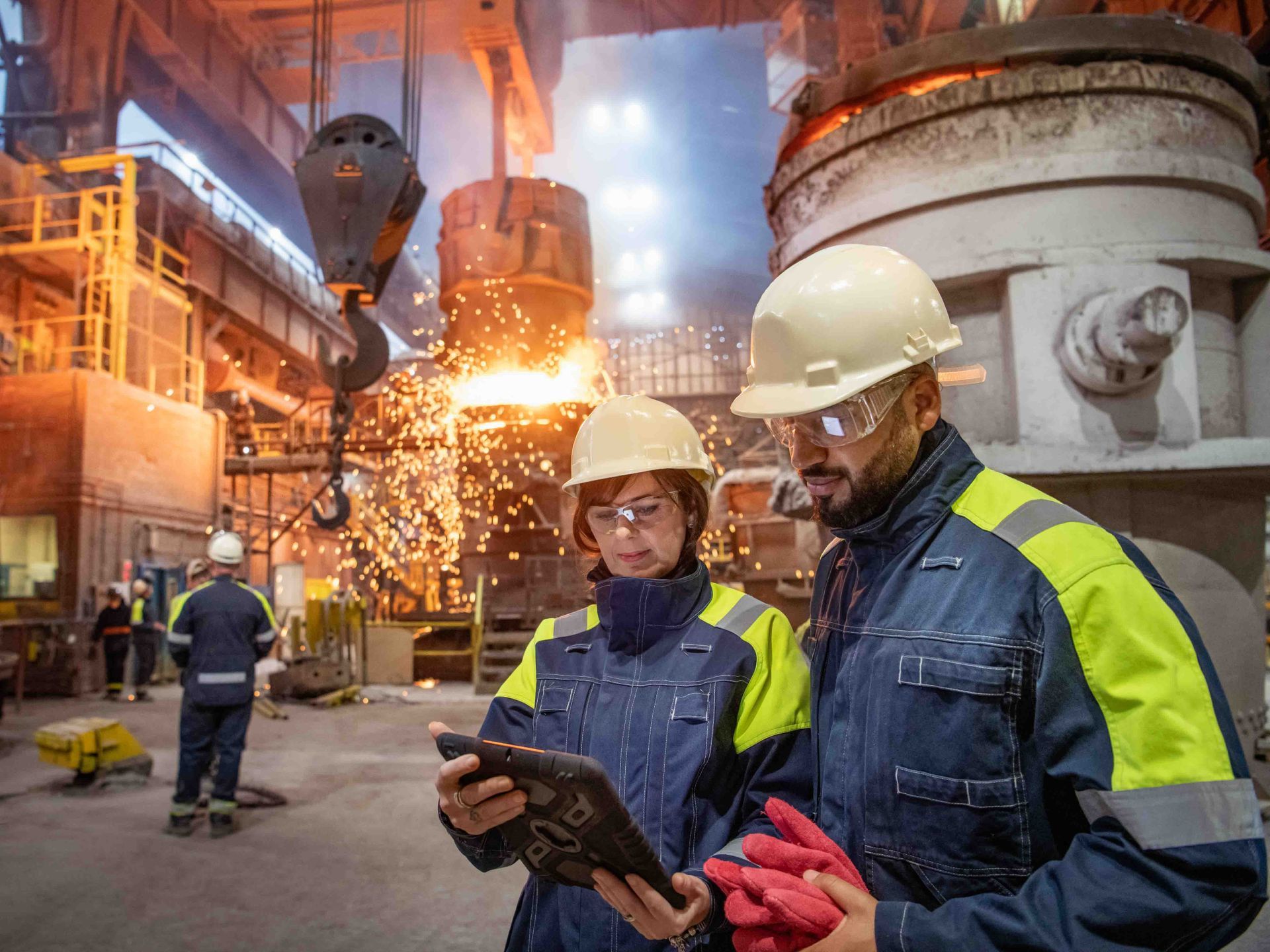 Two people in protective gear examine a tablet in a steel factory with molten metal pouring.