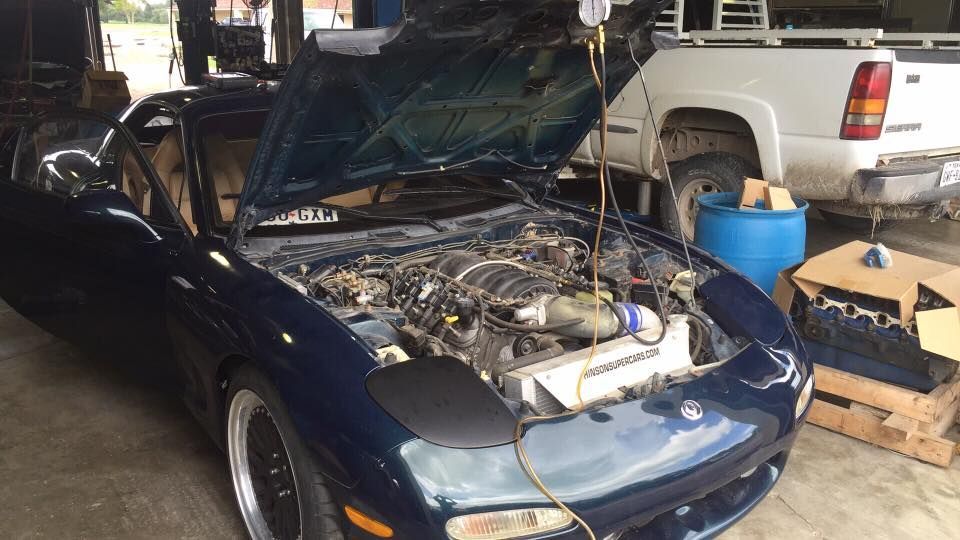 Dark blue car with hood open, exposing the engine, parked in a garage beside a white vehicle.