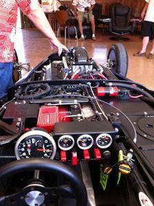 Open race car cockpit with steering wheel, gauges, and exposed engine in a garage.