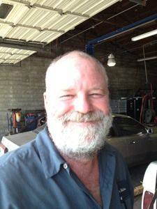 Smiling bearded man in a blue shirt standing in a workshop under a metal roof.