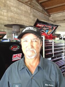 Smiling man in a black cap and work shirt standing in a garage with a toolbox and racing decals