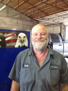 Smiling man in gray work shirt standing in a garage beside a pickup truck and eagle sign.