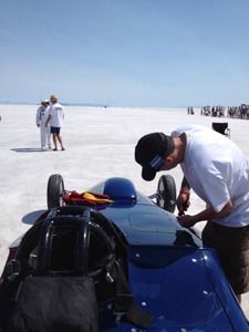 Blue race car and driver being inspected on a salt flat with spectators in the distance