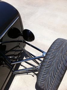 Close-up of a black hot rod’s front suspension and tire, with exposed steering rods and headlights.