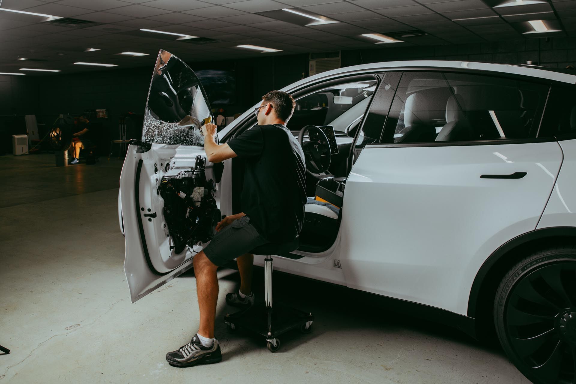 A person tints a white car window. They are inside the open door of a workshop.