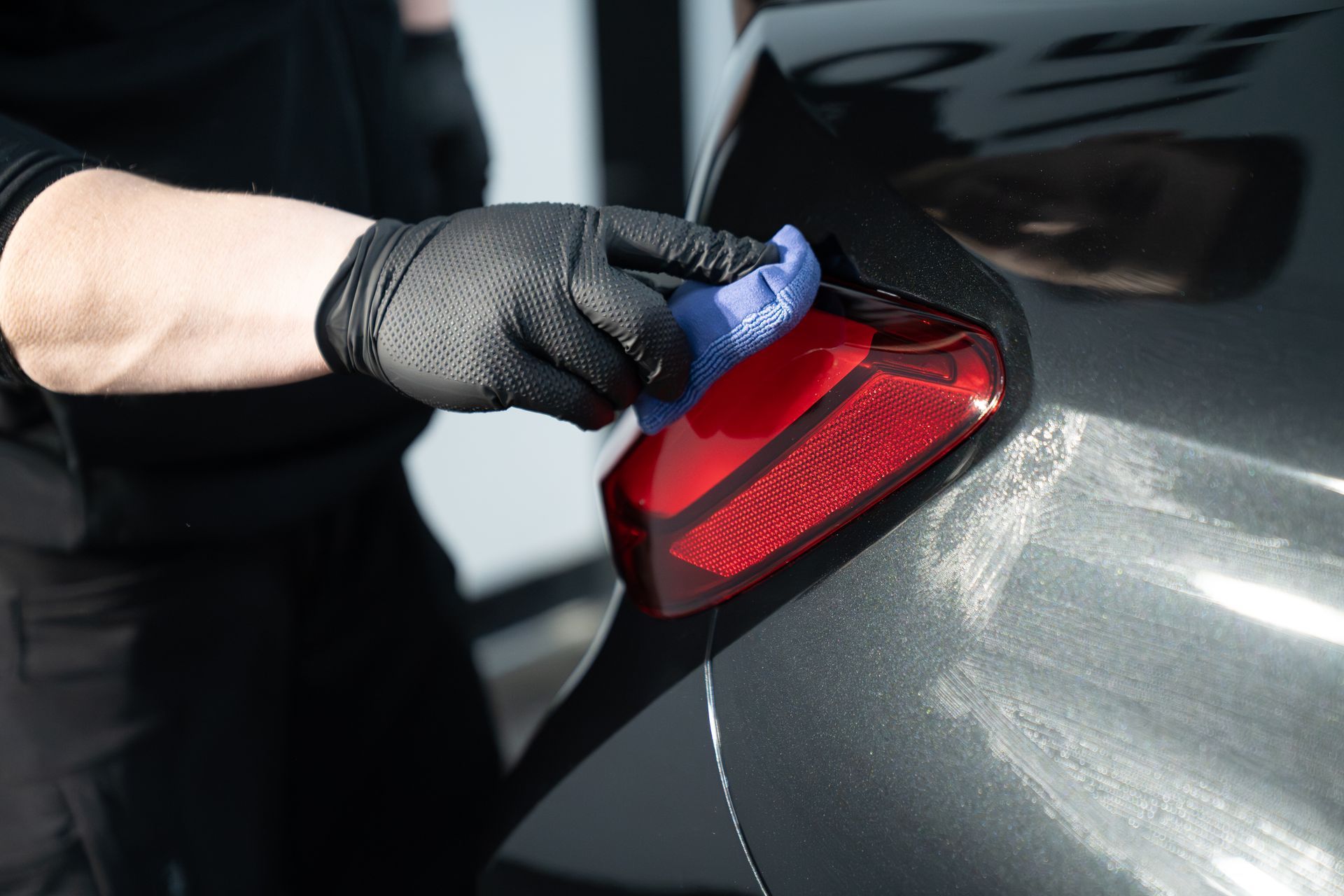 A gloved hand uses a blue cloth to wipe a red vehicle taillight on a dark gray car.
