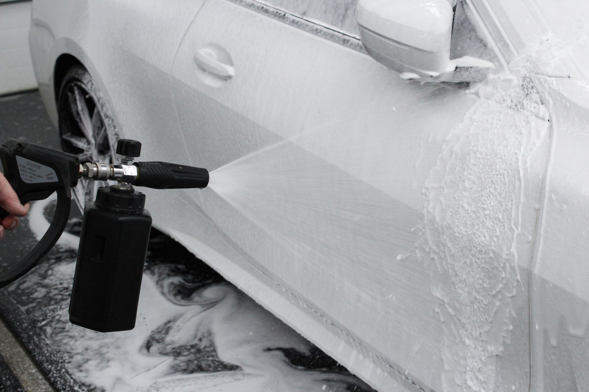 A hand uses a black foam cannon to spray white cleaning soap onto the side of a white car.