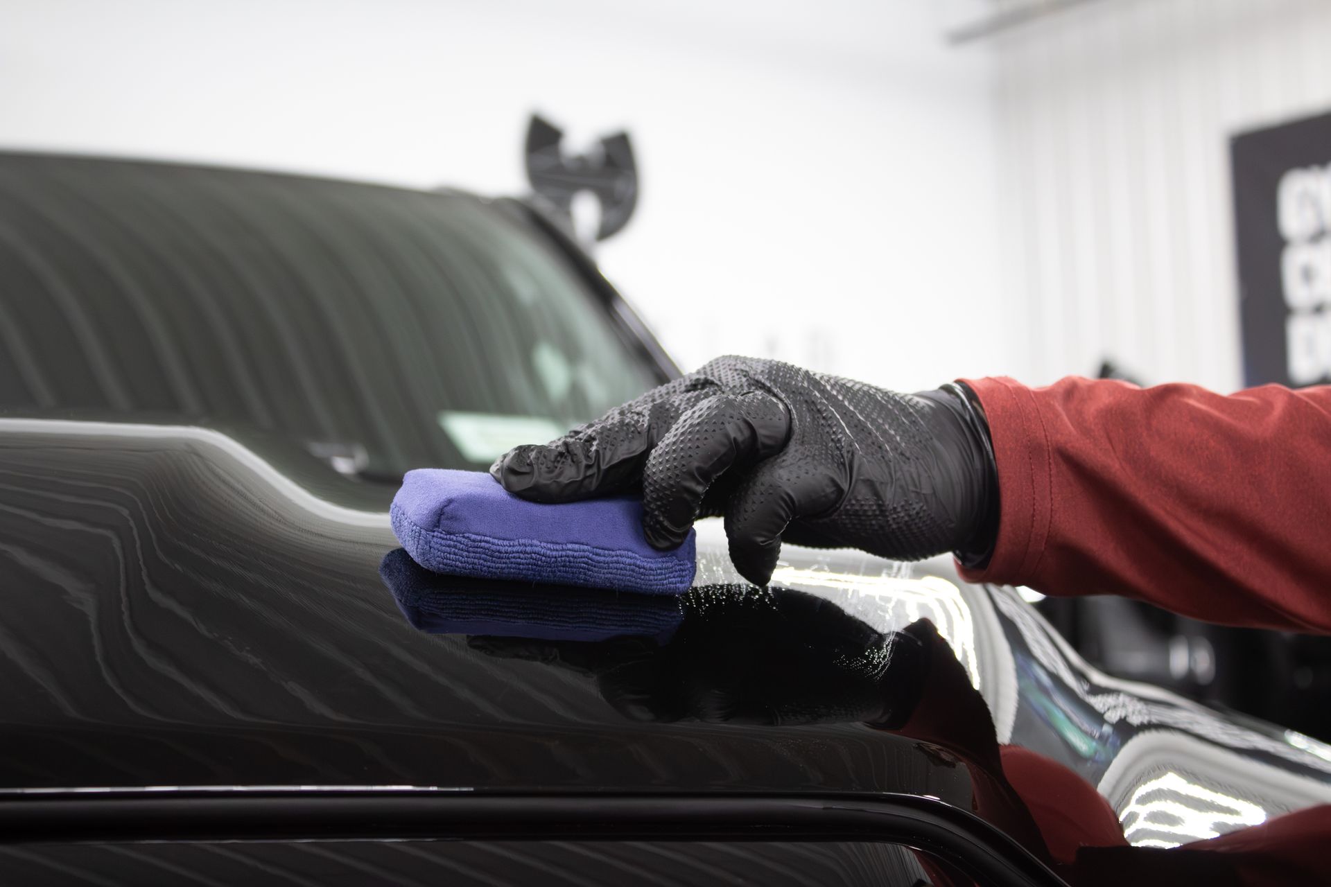 A person wearing a black nitrile glove uses a blue applicator sponge to apply a coating to a shiny black car hood.