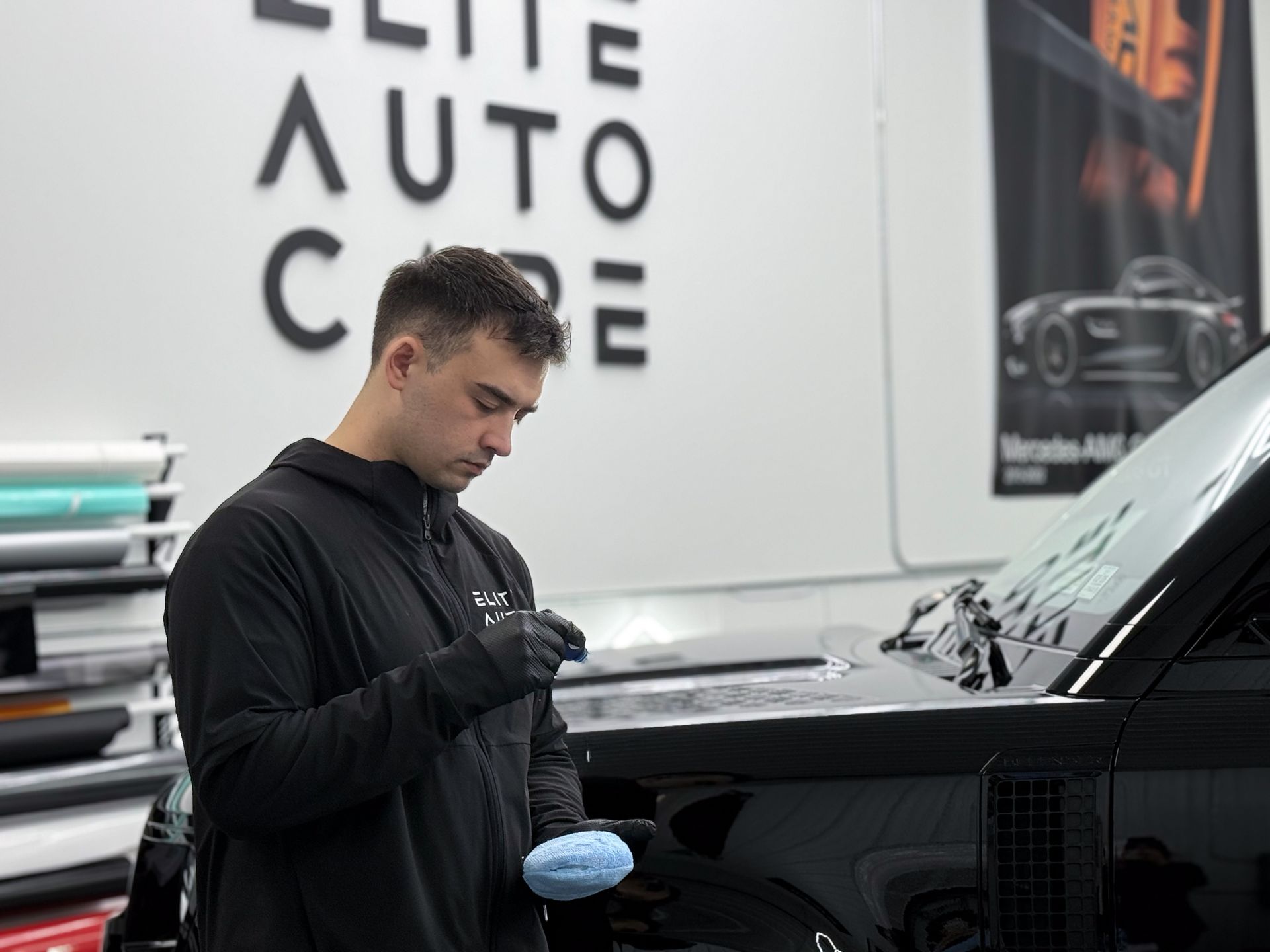A technician in a black jacket and gloves holds a blue detailing pad while preparing to work on a black vehicle.