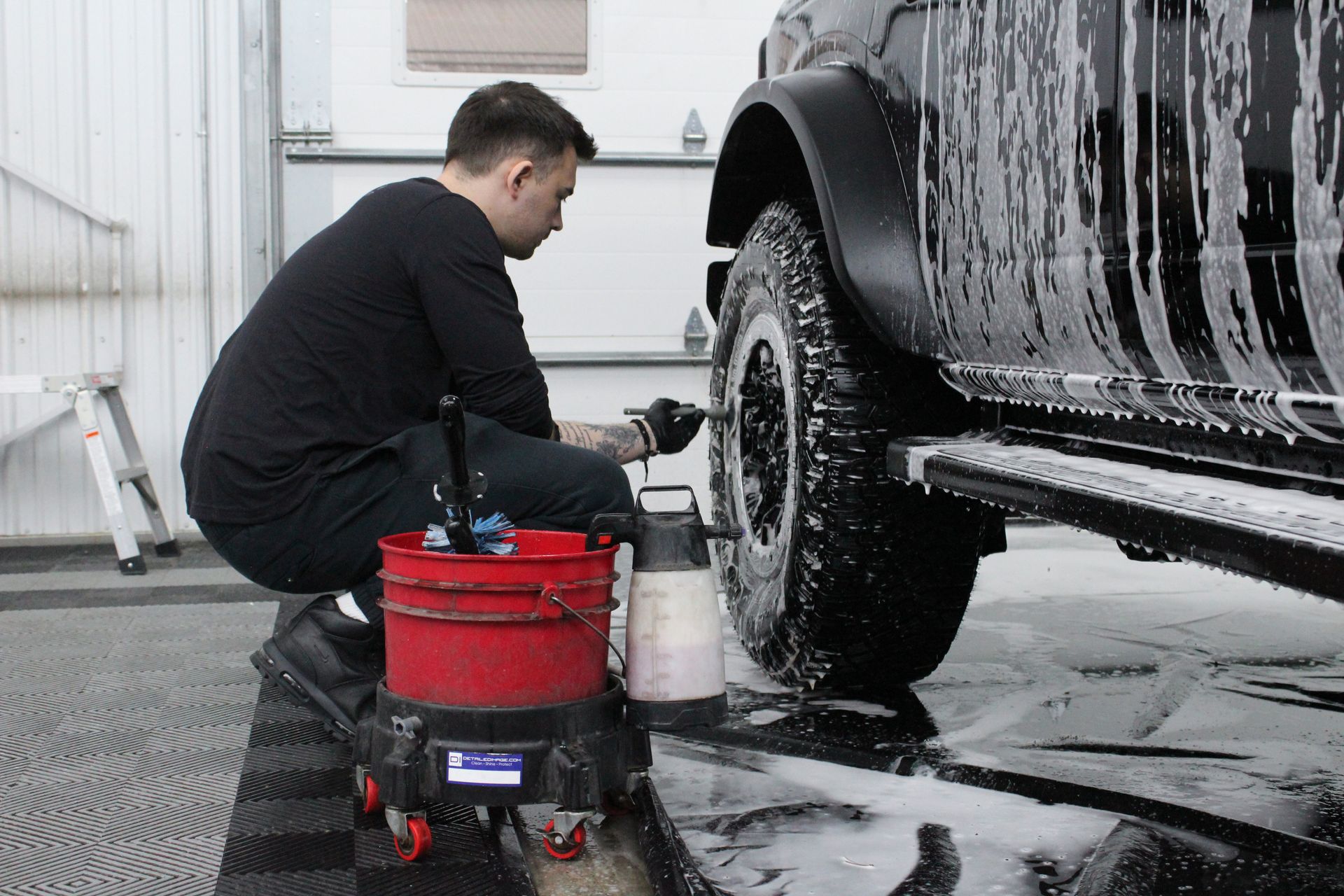 A person in black clothing kneeling to scrub the tire of a foamy, soap-covered vehicle in a workspace.