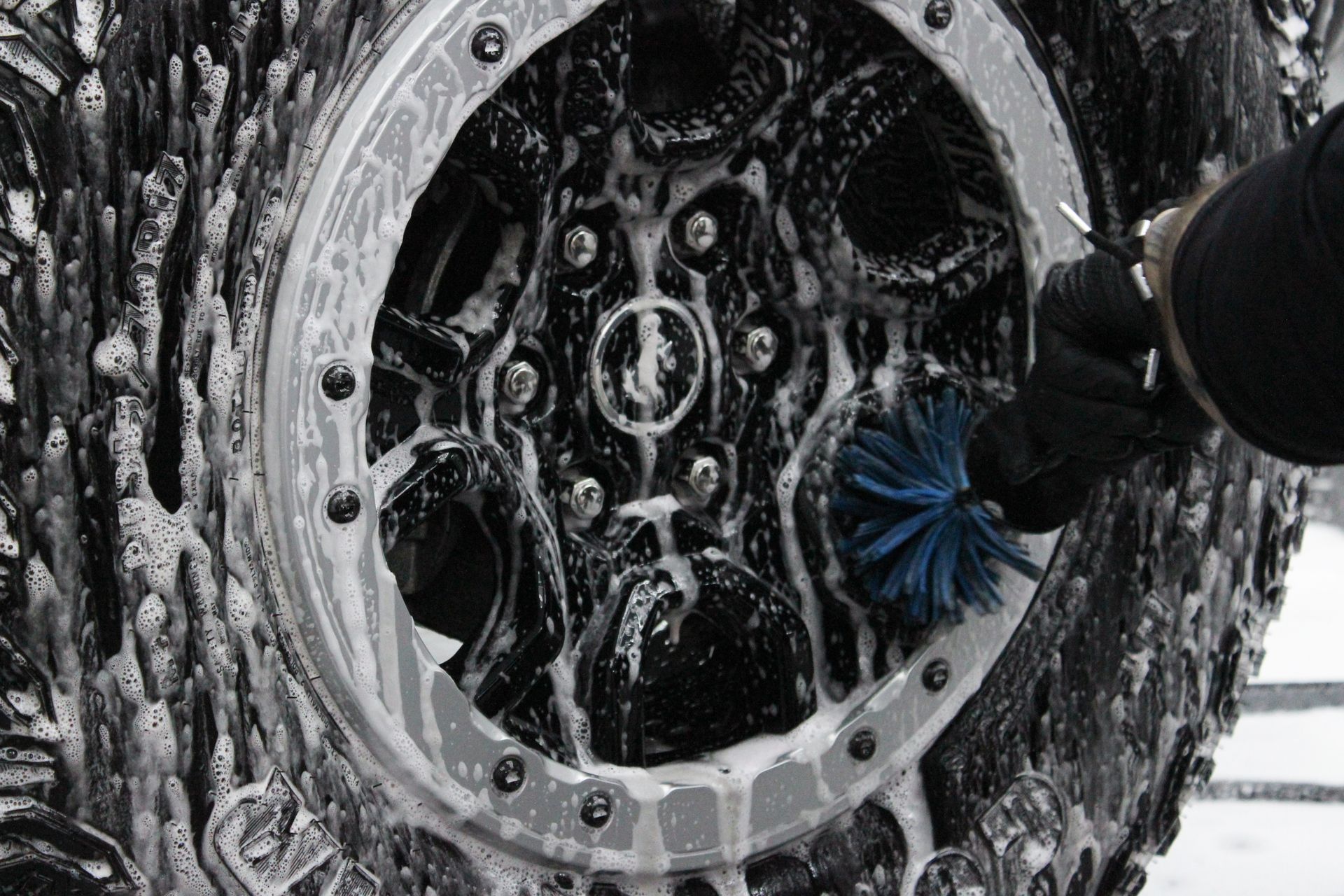 A person using a blue brush to scrub a soapy, mud-caked vehicle tire and wheel.