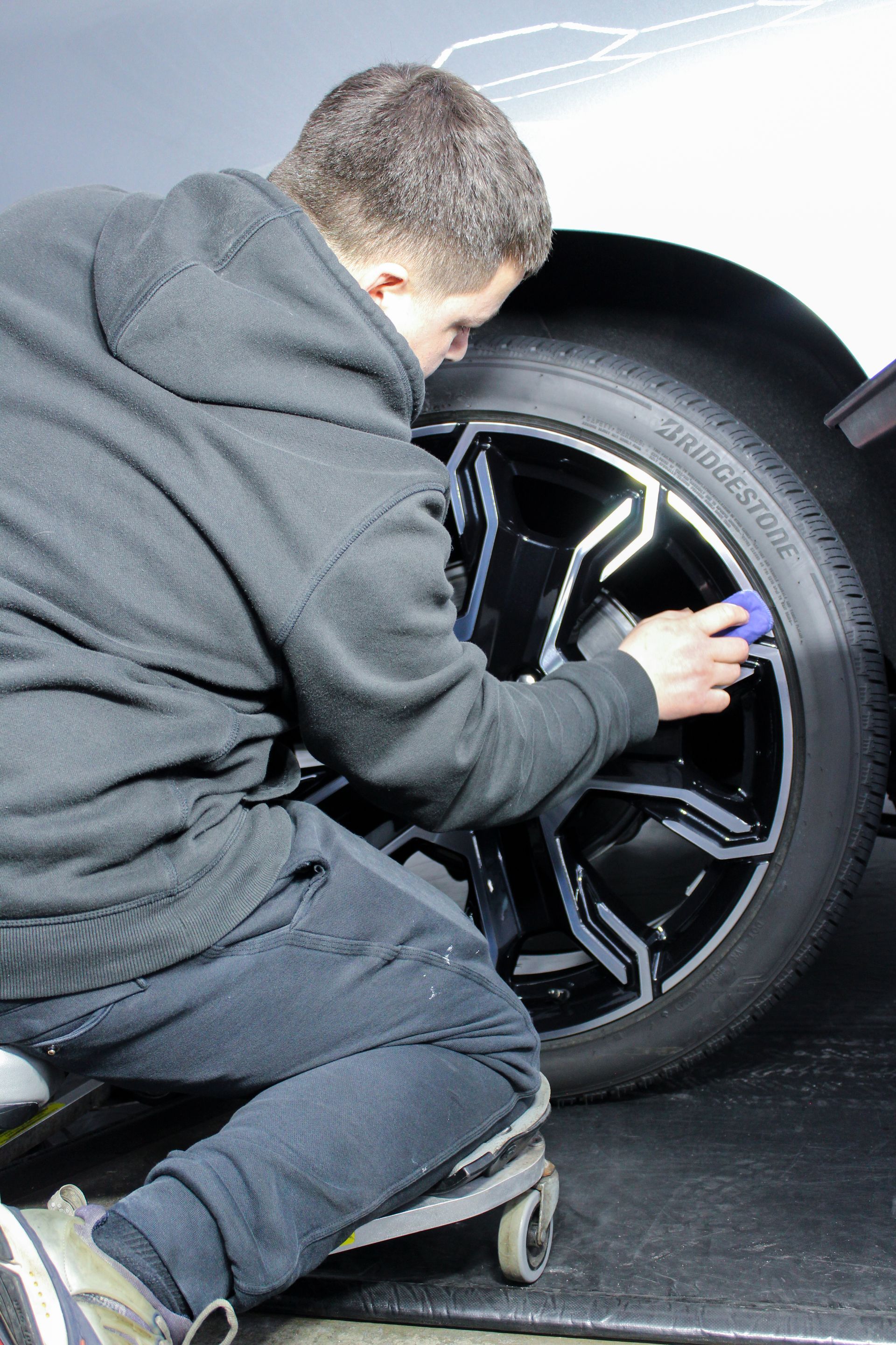 A person wearing a grey hoodie kneels on a wheeled seat, applying product to a black car wheel with a small applicator.