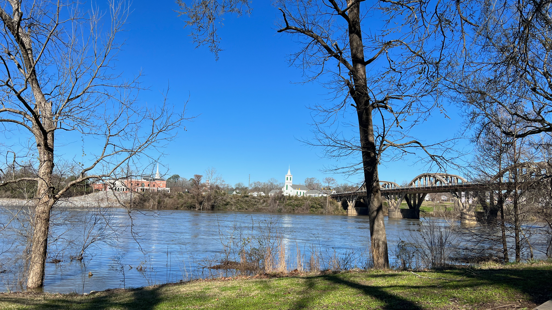 River view with bridge, town, trees, and blue sky.