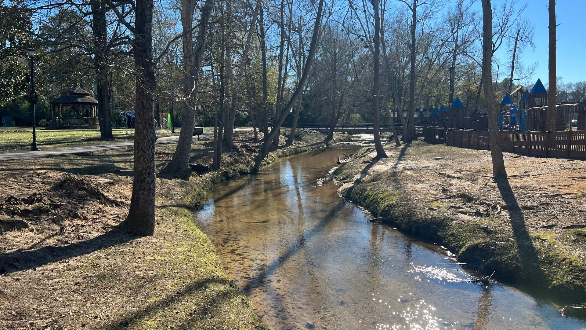 Stream flowing through a park, lined with trees and a playground in the background.