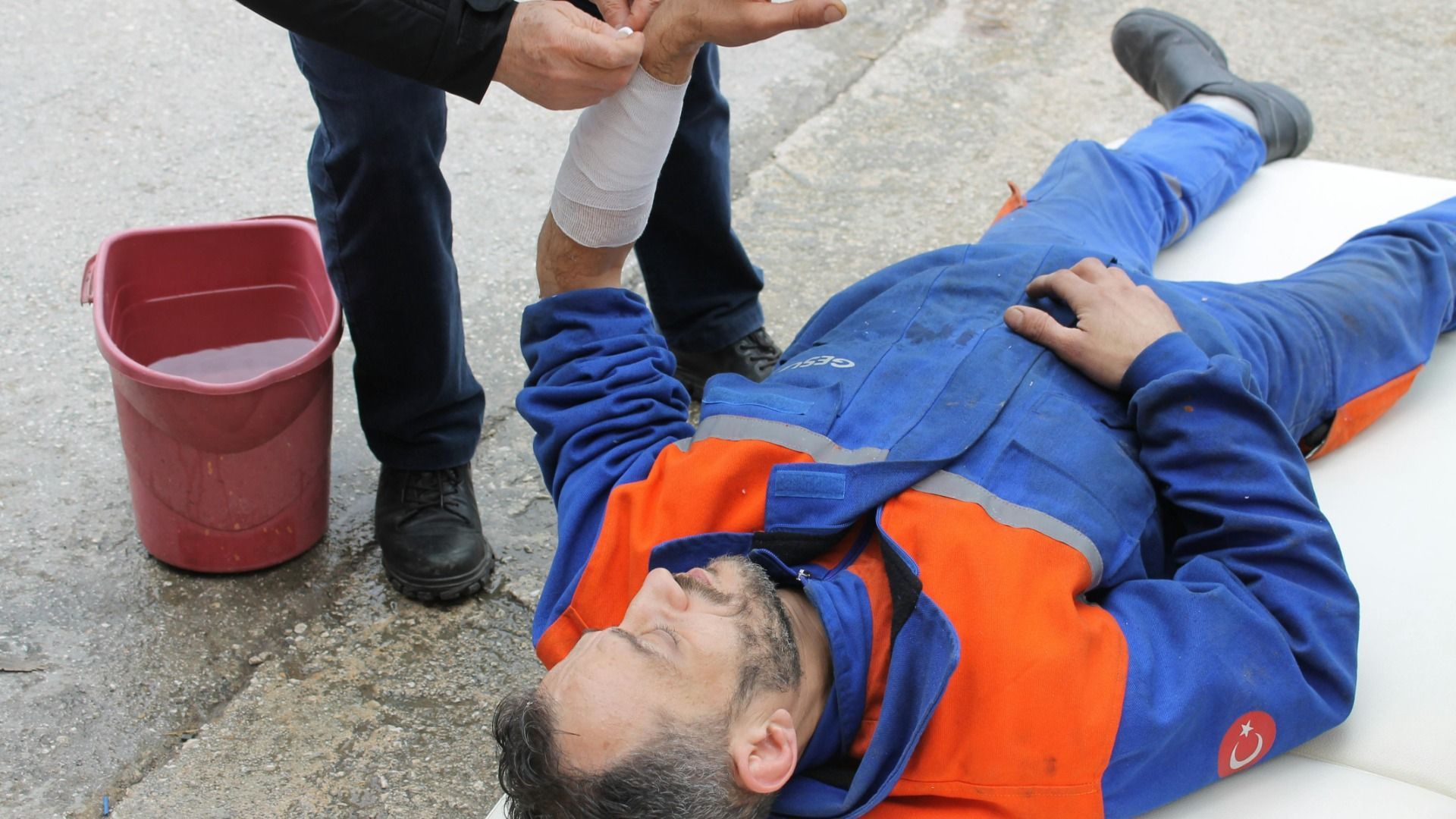Person bandaging another person's arm, who is lying down on a white surface, a bucket of water beside them.