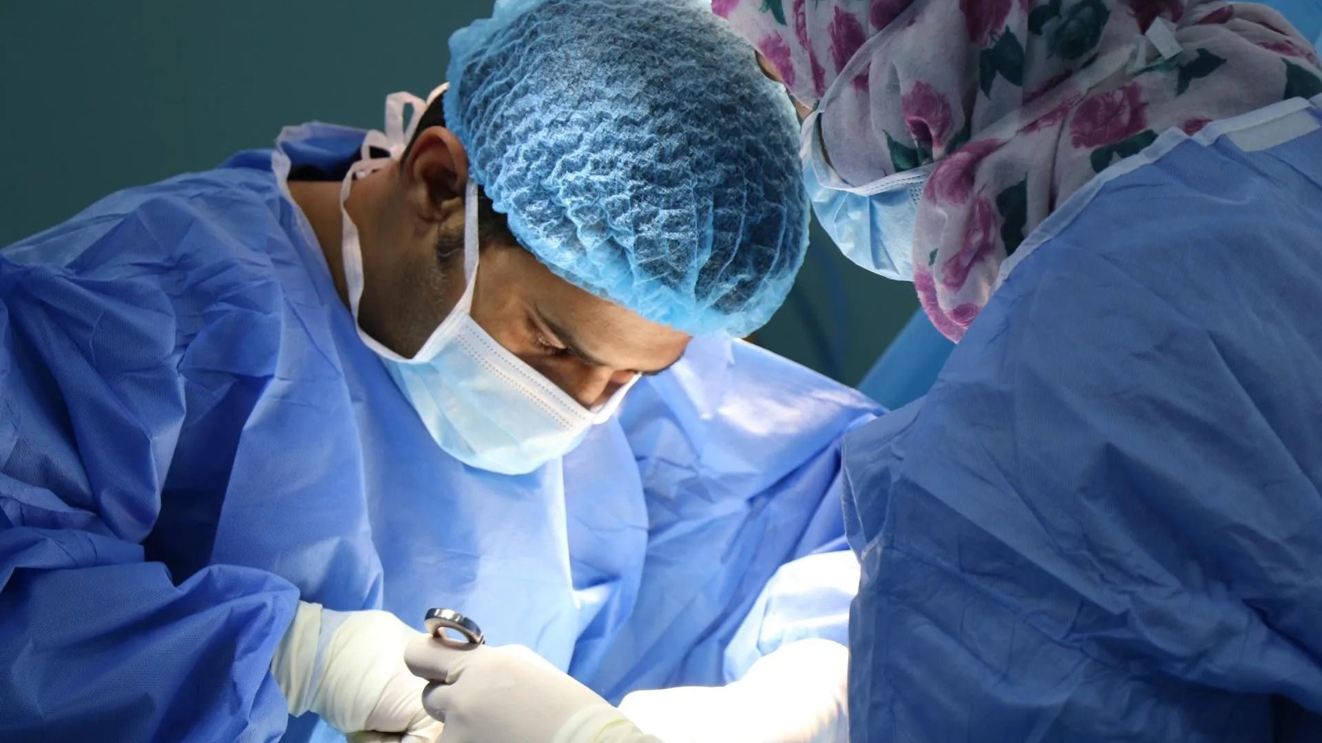 Surgeons in blue scrubs and caps perform surgery, focused, illuminated, in an operating room.