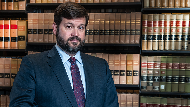 Man in suit with beard, standing in front of law books, looking at the viewer.