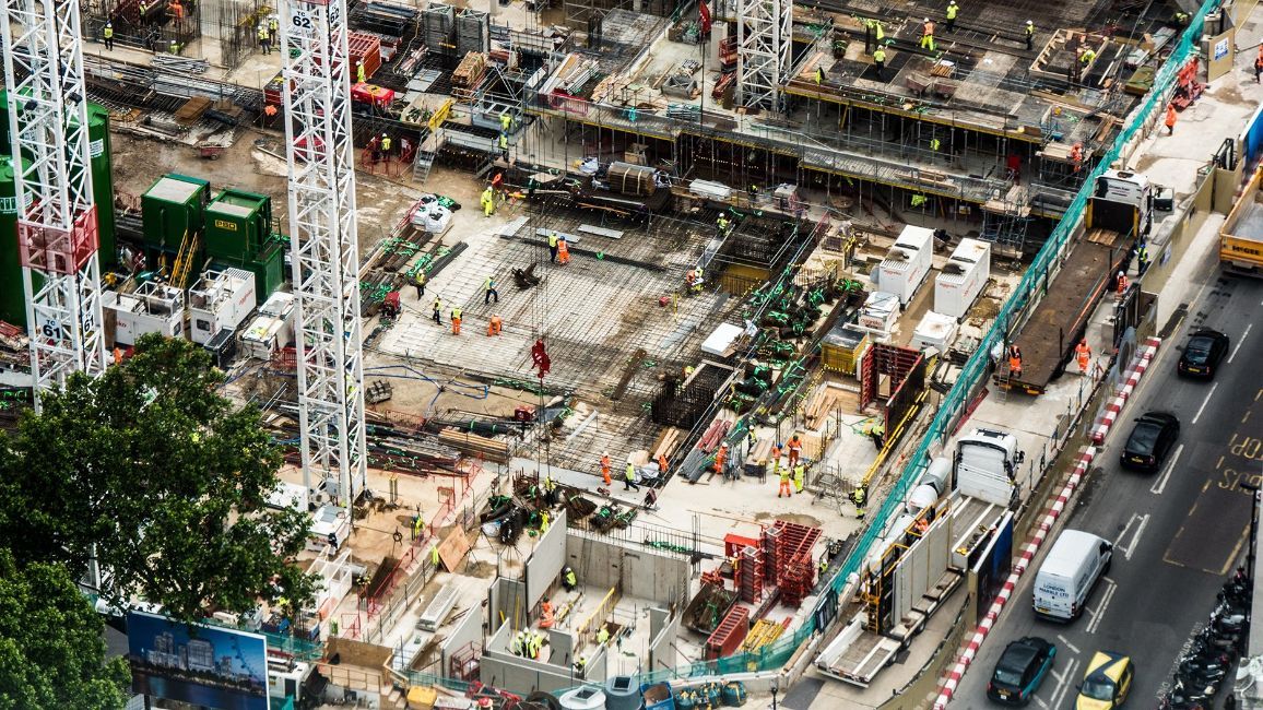 Construction site with cranes, workers, and materials; next to a street with traffic.