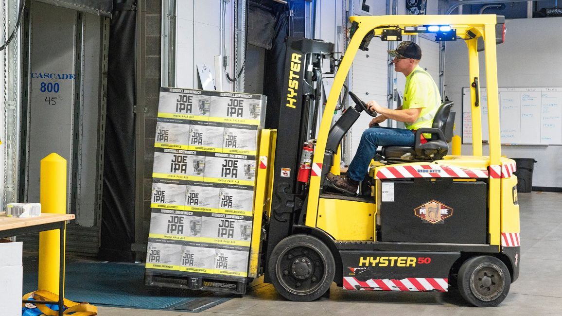 Forklift operator moving stacked boxes inside a warehouse. Yellow forklift; worker wearing a yellow vest.