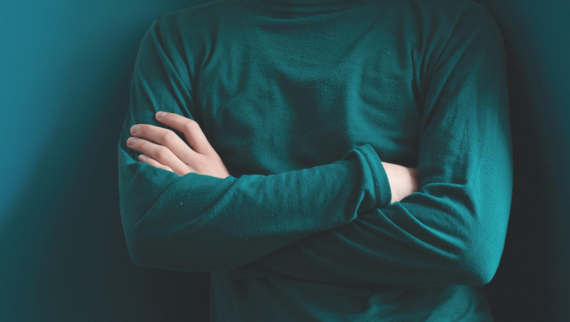 Person in teal sweater, arms crossed, leaning against a teal-colored wall in a corner.