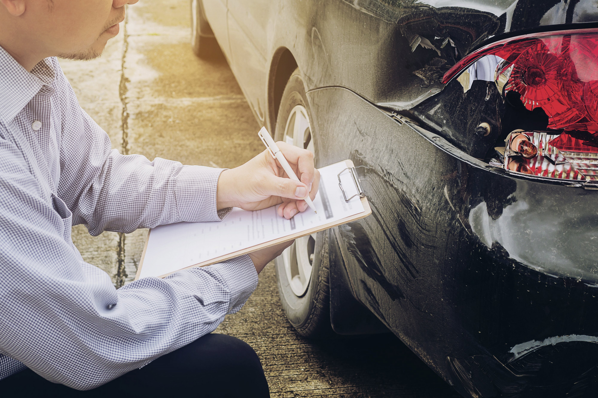 Insurance adjuster inspecting and documenting rear-end collision damage on a black car with a clipbo