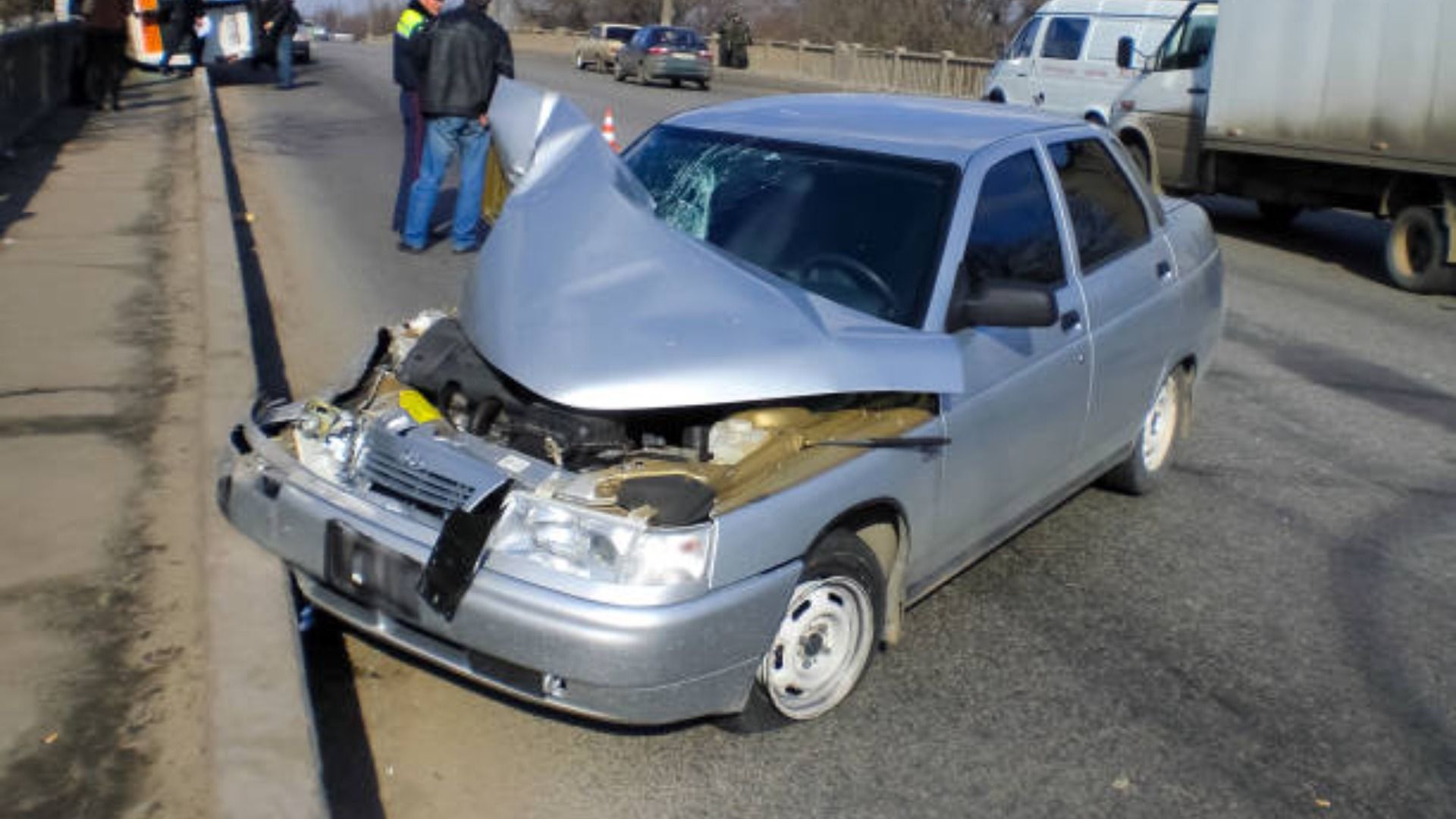 Silver car damaged in a traffic accident on a bridge; emergency services personnel visible.