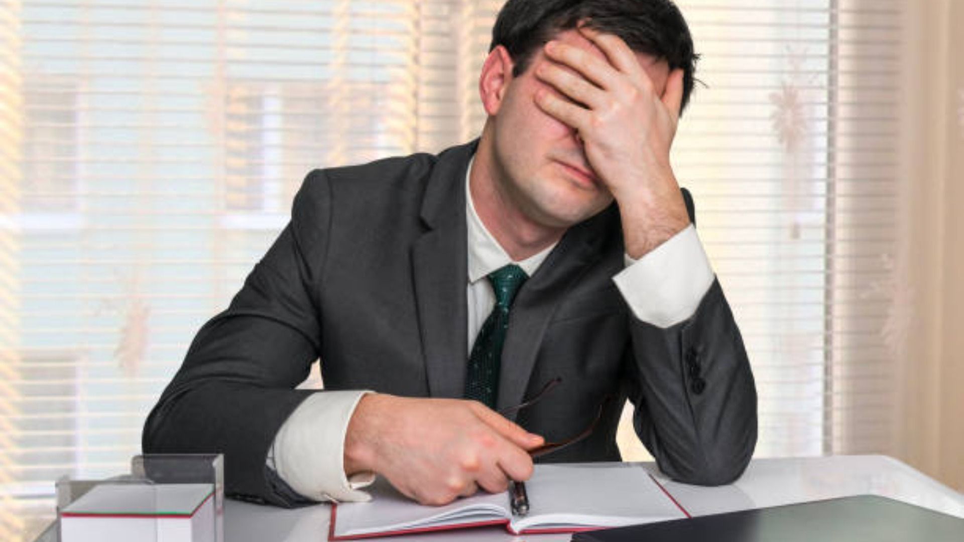 Man in suit, hand over face, appears stressed, sitting at a desk.