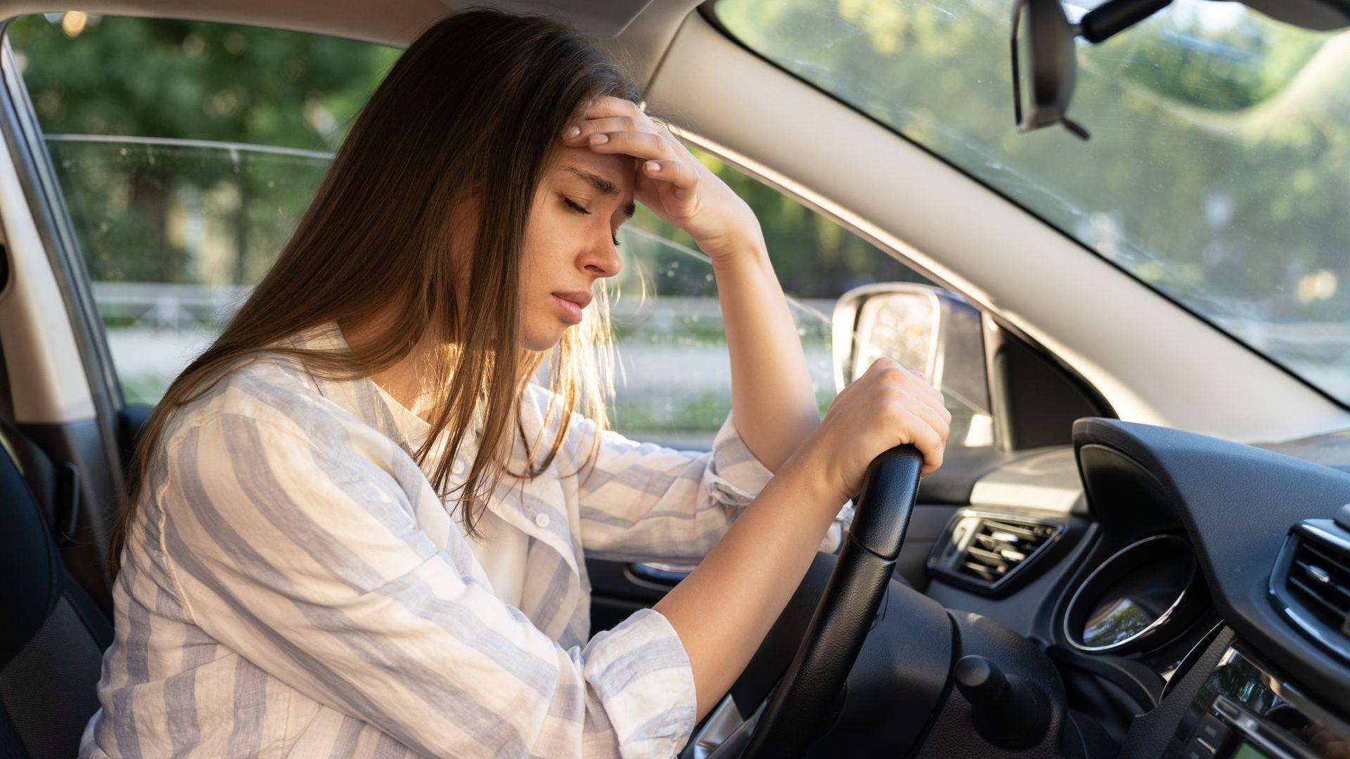 Woman in a car, hand on forehead, looking stressed. Sunlight streams in.
