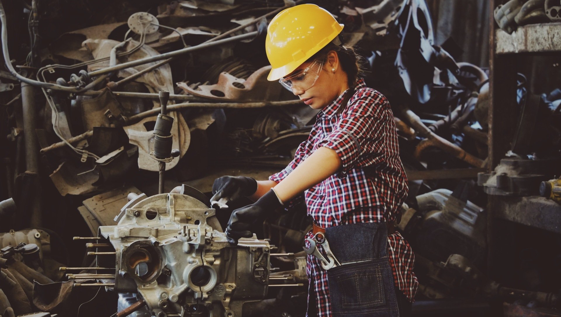 Woman in yellow hard hat and safety glasses works on machinery in a cluttered workshop.