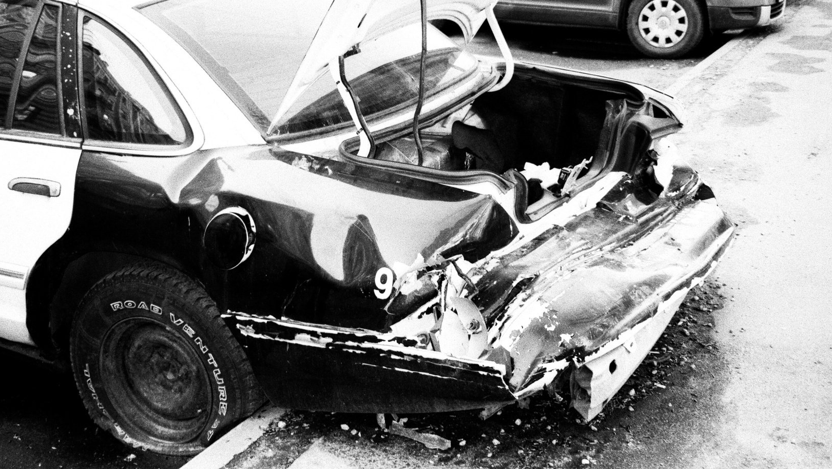 Rear view of a damaged black and white police car with the trunk open, dented from a crash.