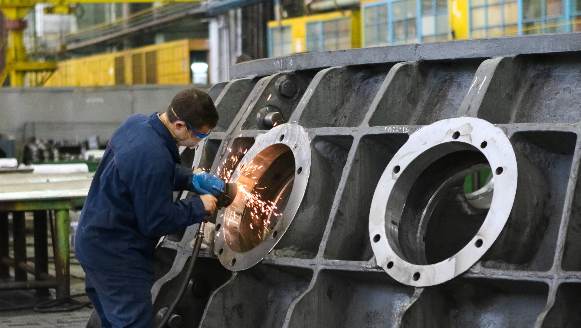 Man in safety glasses grinds metal, creating sparks, in a factory setting.