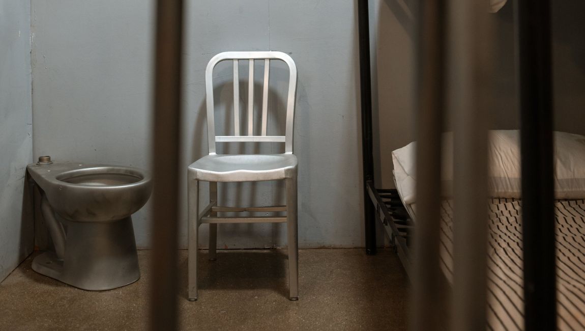 Prison cell with toilet, chair, and bunk bed; viewed through bars.
