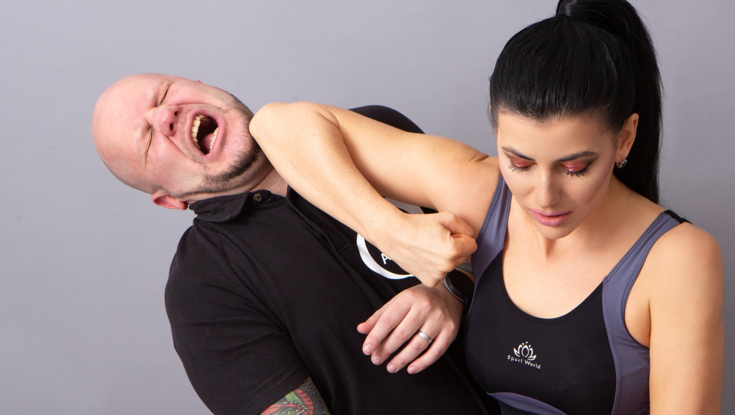 Woman applying arm bar to man's neck; man grimaces; gray backdrop.