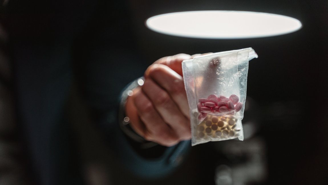 Hand holding a clear bag with pink and yellow pills, under a bright light.