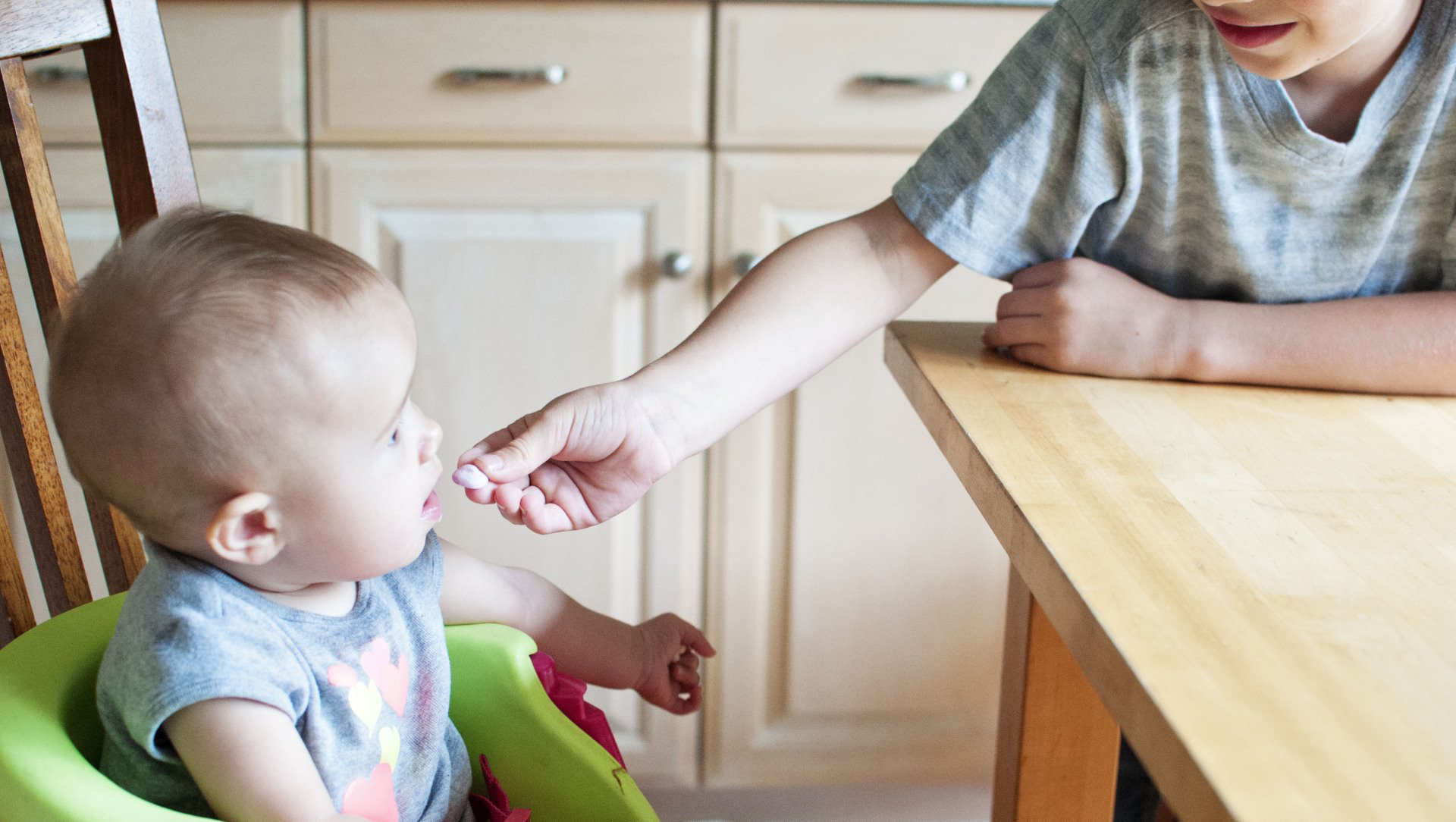 Child feeding a baby in a high chair at a wooden table. Kitchen cabinets in the background.