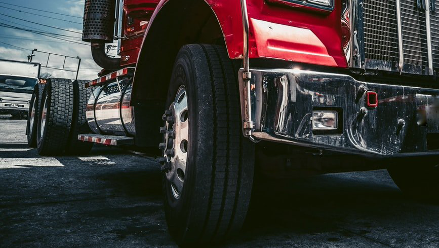 Red semi-truck's front wheels and chrome bumper on asphalt, cloudy sky in background.