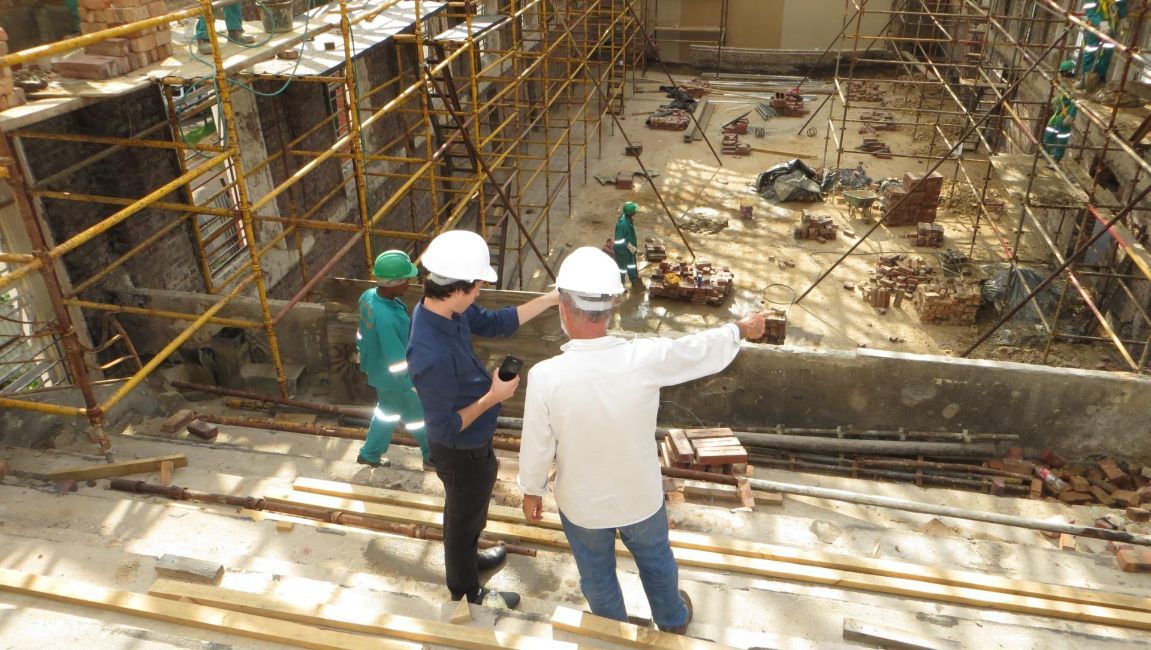 Construction site: Two men in hard hats survey work, pointing. Scaffolding, workers in green, and debris are visible.
