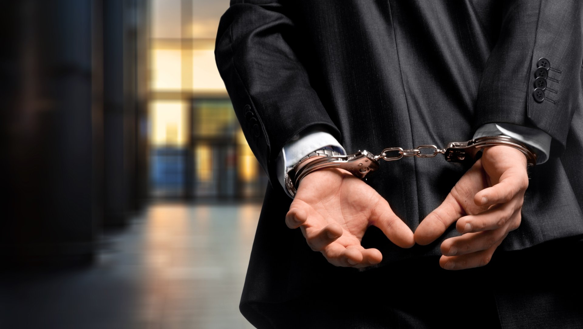 Person in a suit with hands cuffed behind their back. Interior, blurred background.