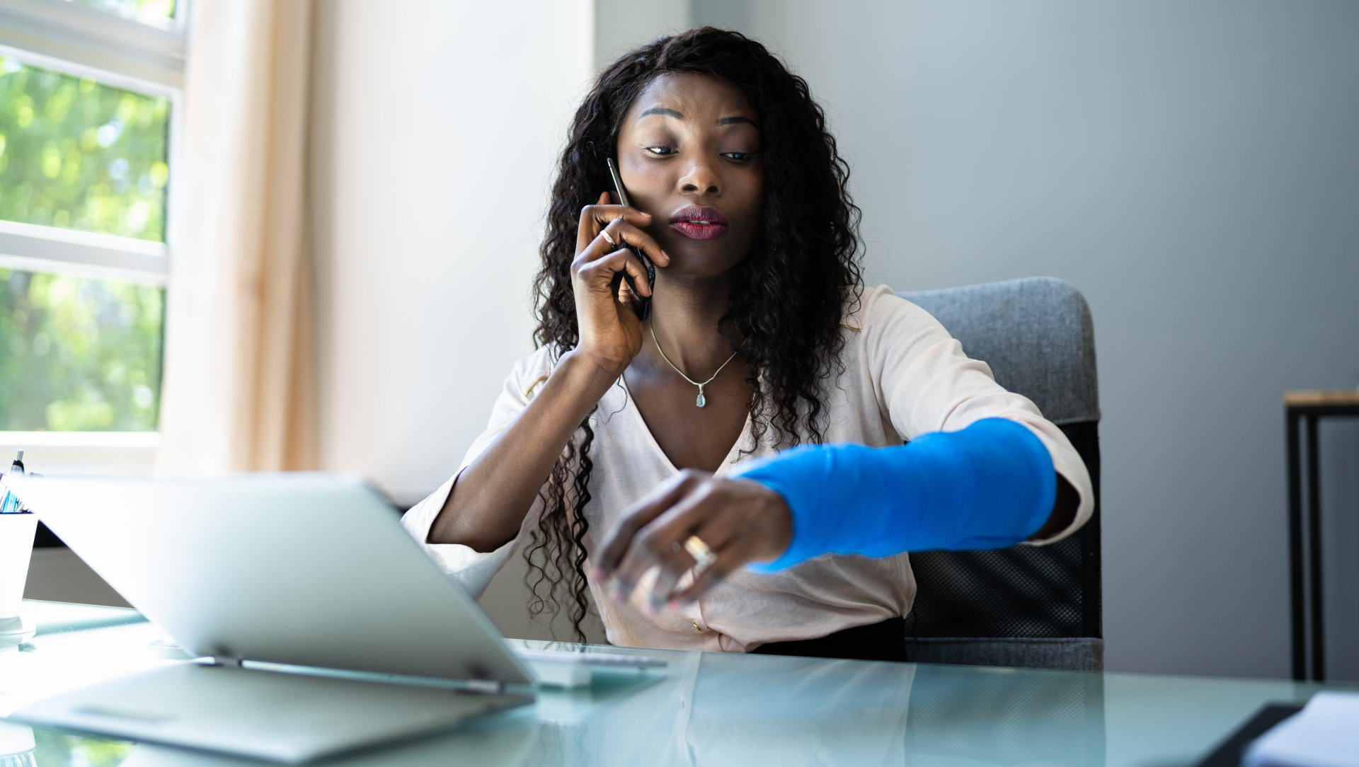 Woman in office on phone with blue arm cast, looking at watch.