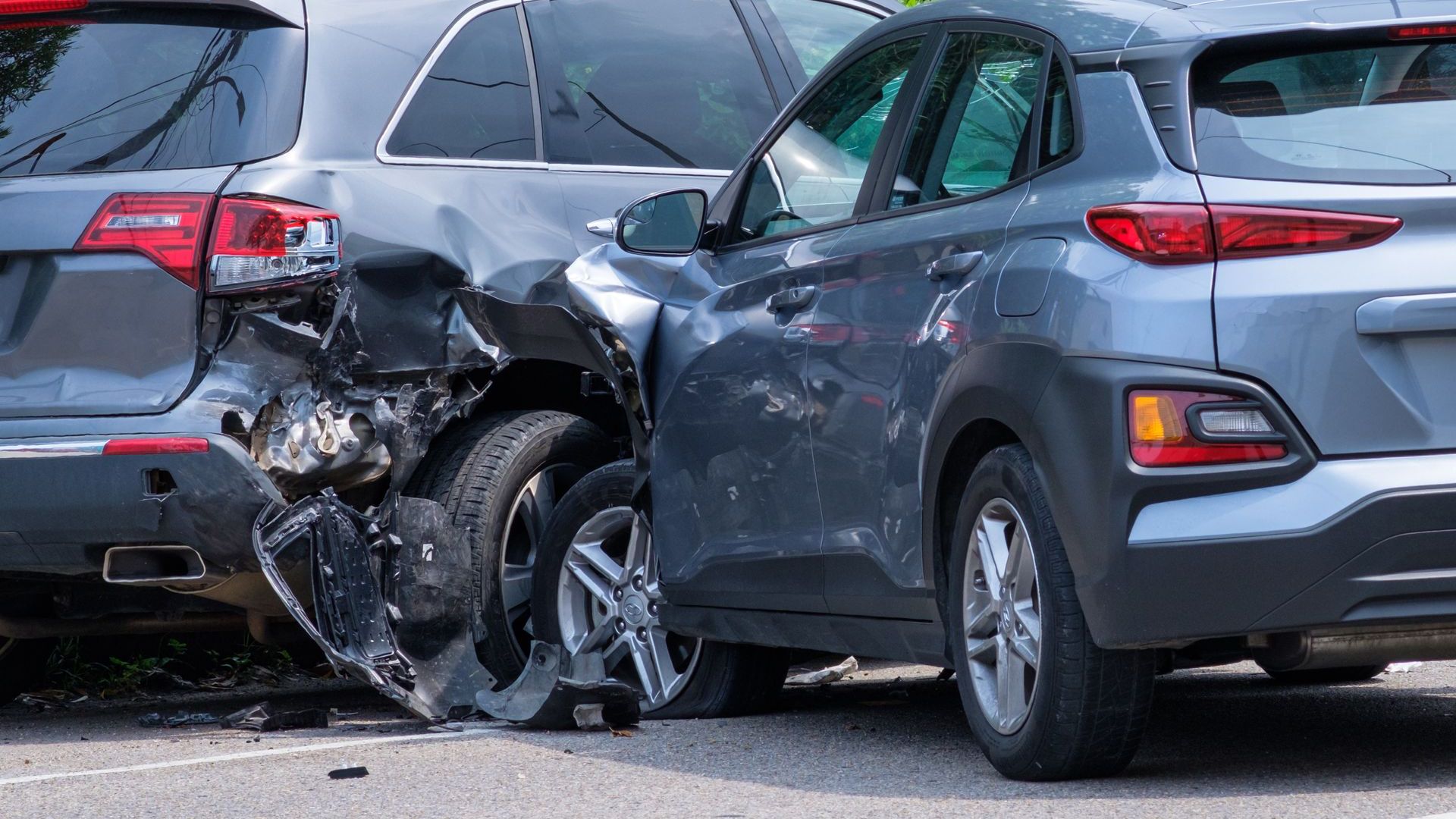 Two gray cars involved in a collision, one with severe rear-end damage, blocking a street.